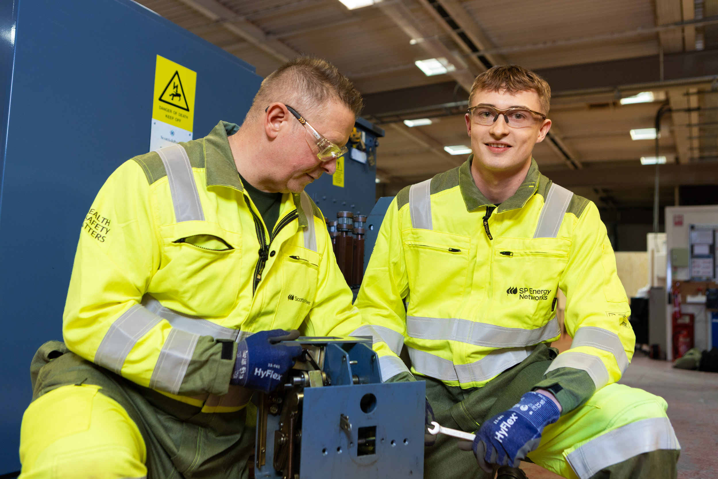 Two men kneel down next to a mechanical part. They're both wearing hi-vis boiler suits and one of them is holding a spanner.