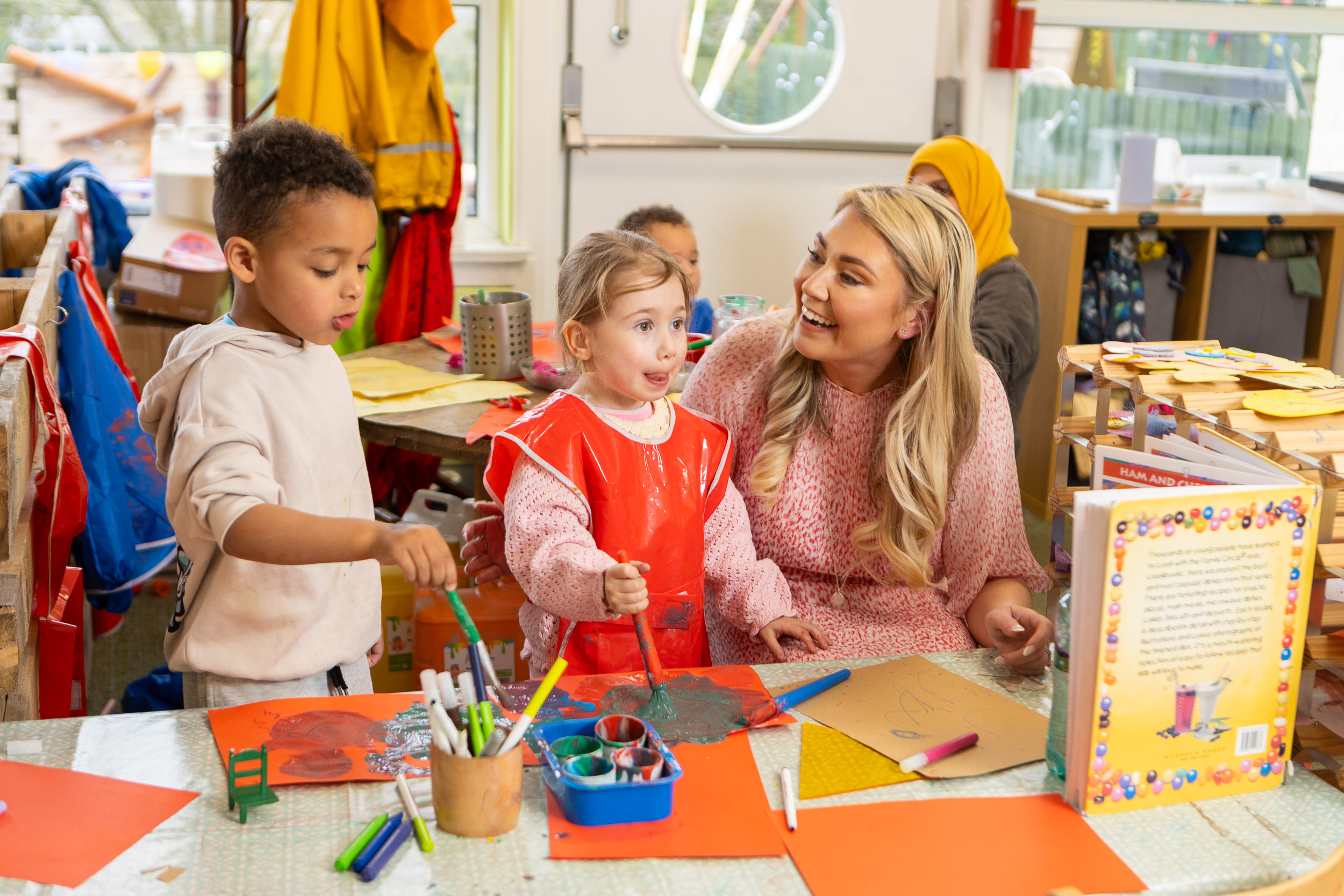 A young woman stands next to two young children in a nursery environment.
