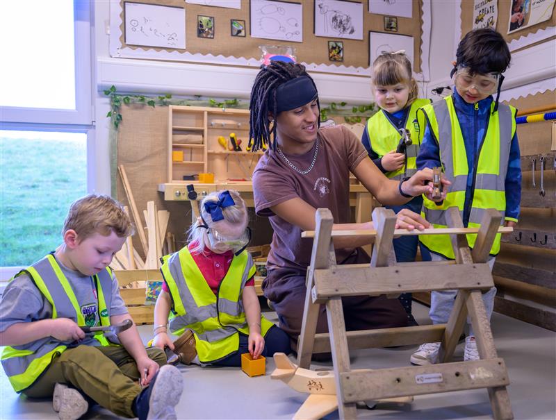 A young man assembles a wooden structure while several children, all wearing hi-vis vests, stand next to him.