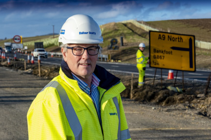 A man standing at the side of a road wearing a hardhat and a high visibility jacket.