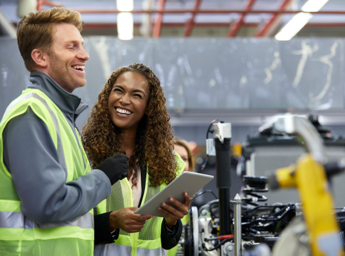 A man and woman, both wearing high visibility vests, looking at each other and laughing. The woman is holding a notepad.