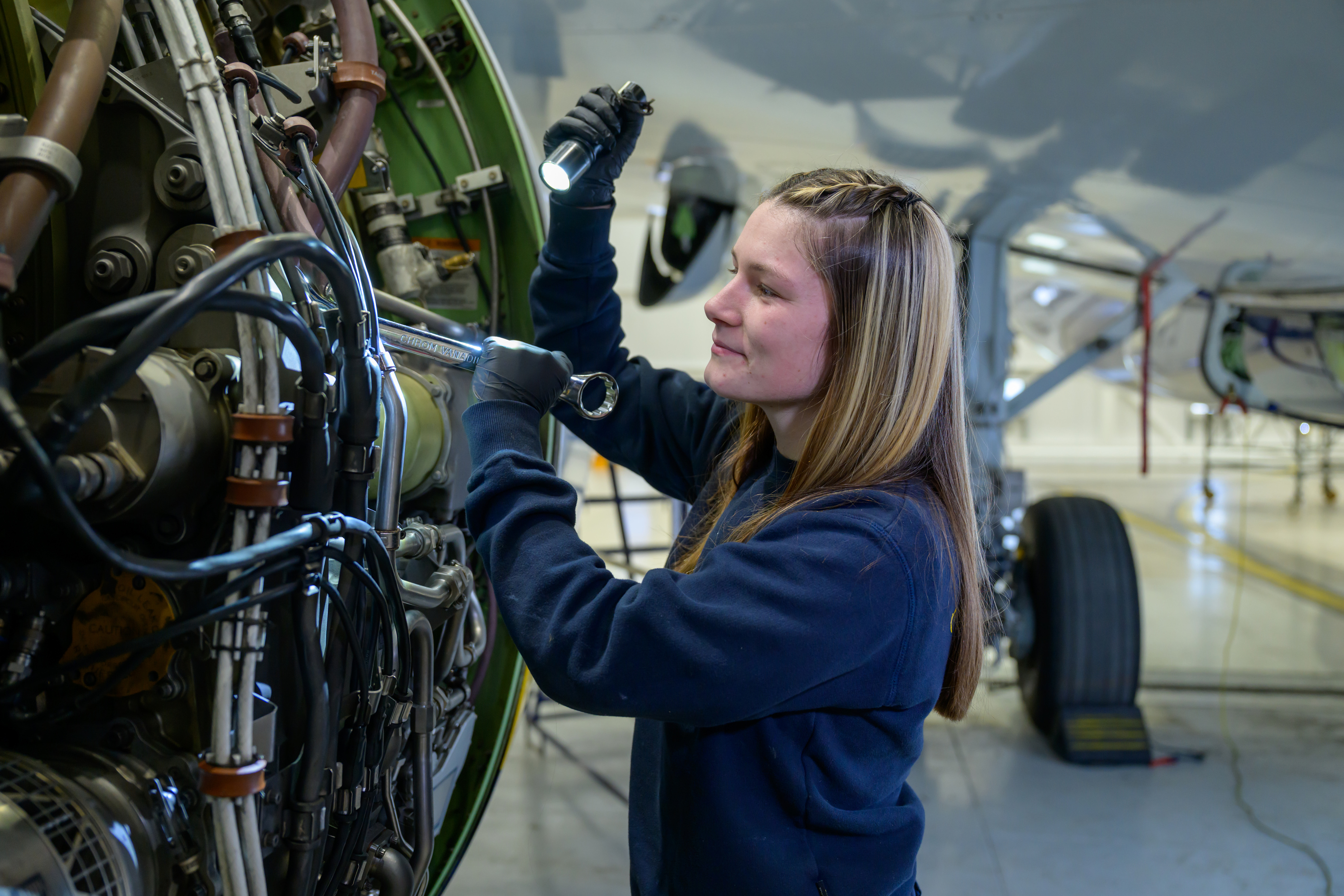 A young woman, wearing a boiler suit, works on a section of aircraft engine.
