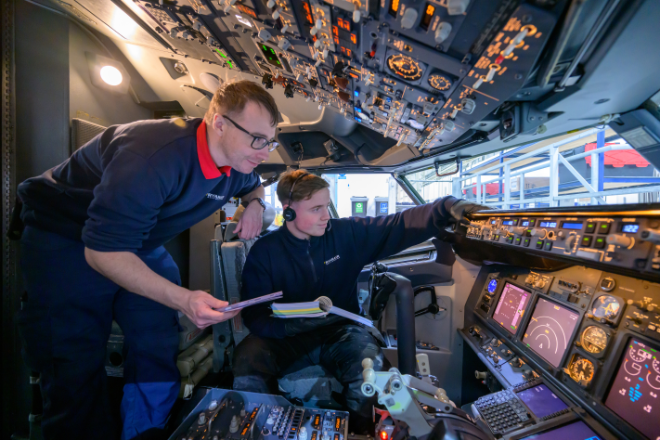 A young man sits in the cockpit of a plane while another man stands behind him and holds a notepad.