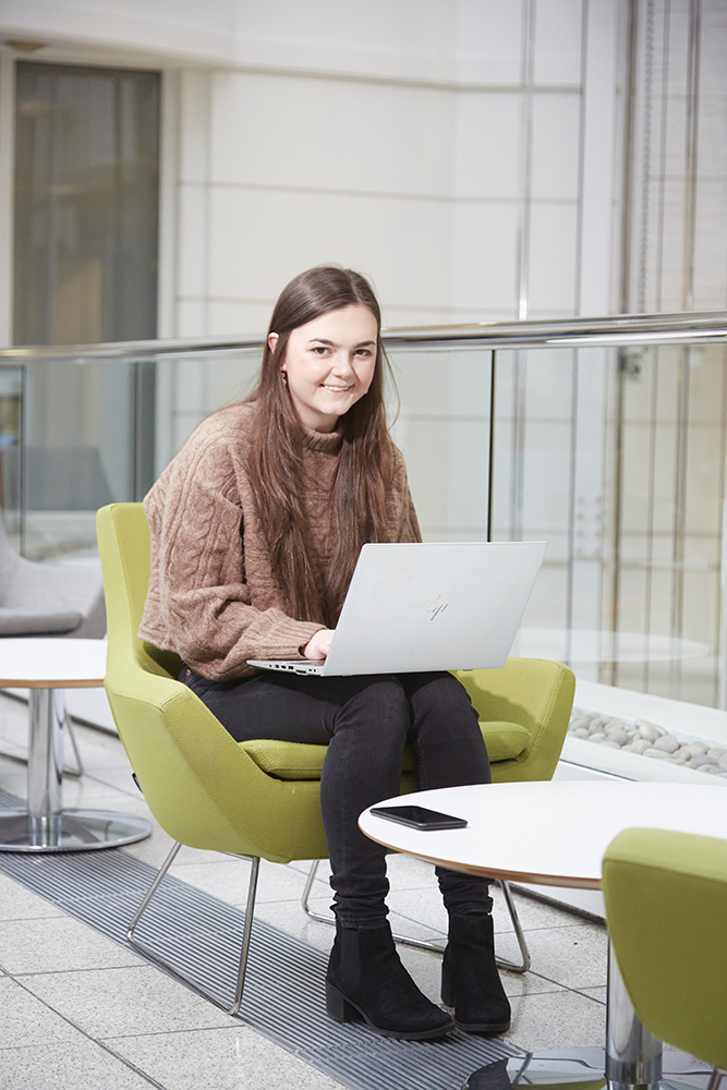 A woman smiling at the camera while using a laptop which is sitting on her knees.