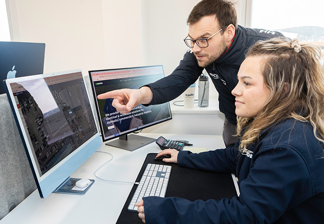 A woman sitting down using a computer while a man points out something to her on the screen.