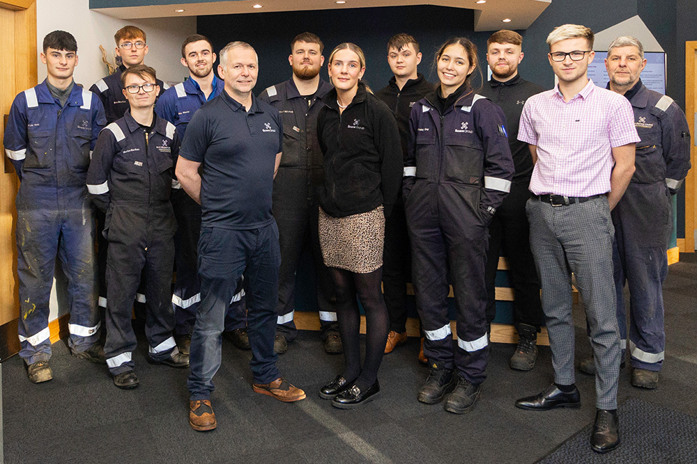 Twelve people, some wearing boiler suits and some wearing officewear, standing at a reception desk.