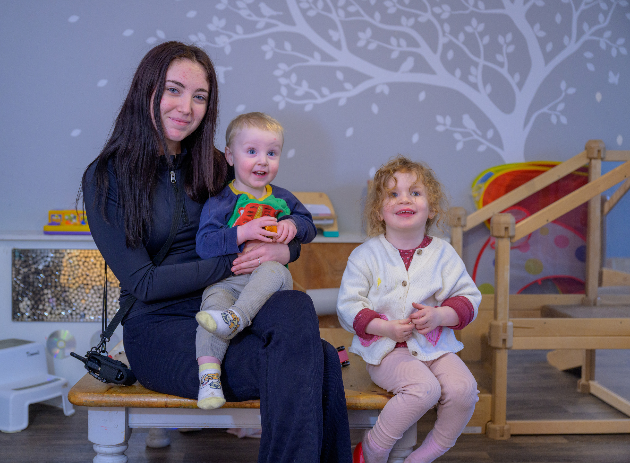 A young woman sits on a bench within a nursery environment with a child on her knee and another child sitting next to her.
