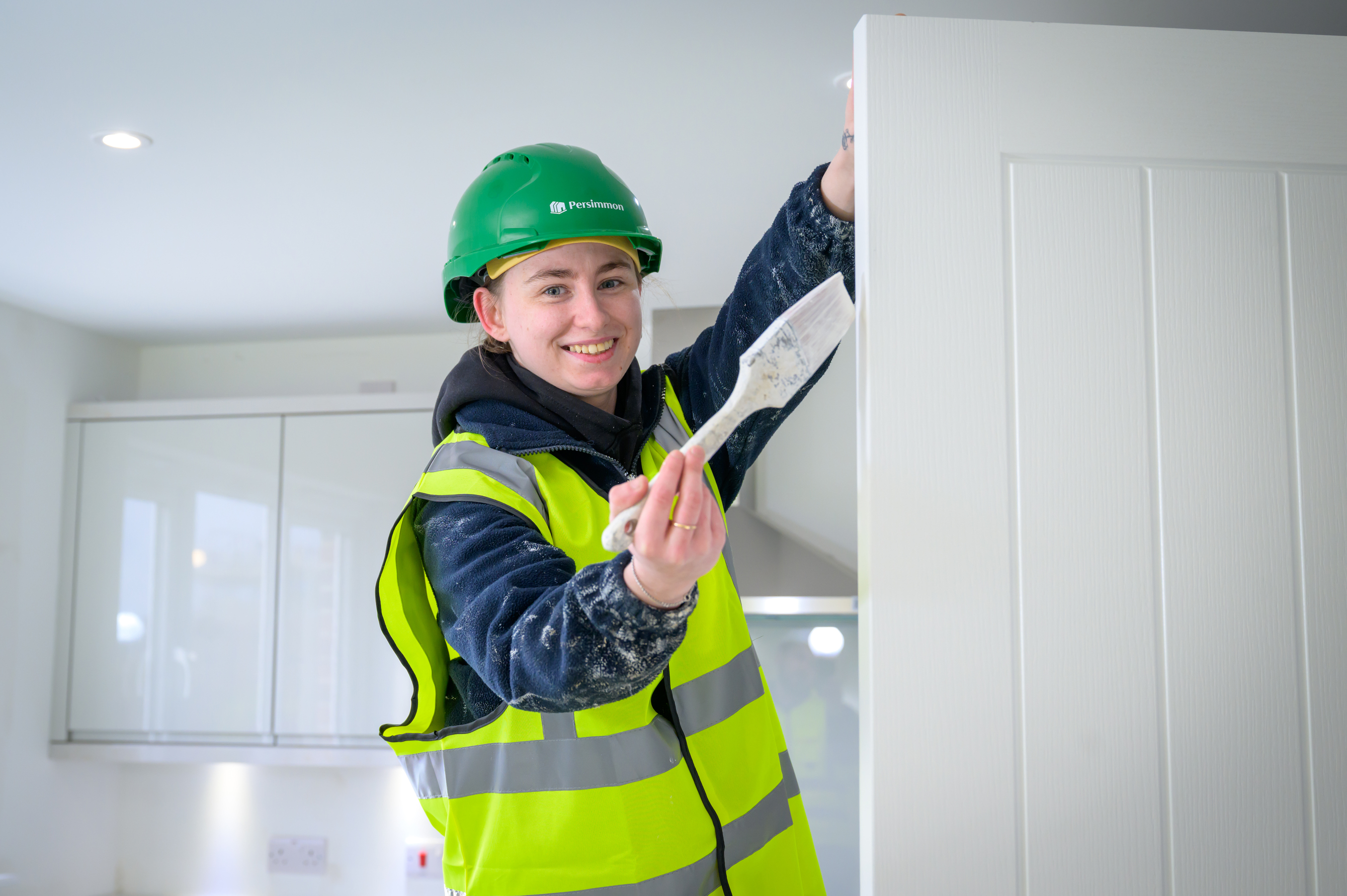 A young woman holds a paintbrush and wears a hi-vis vest.