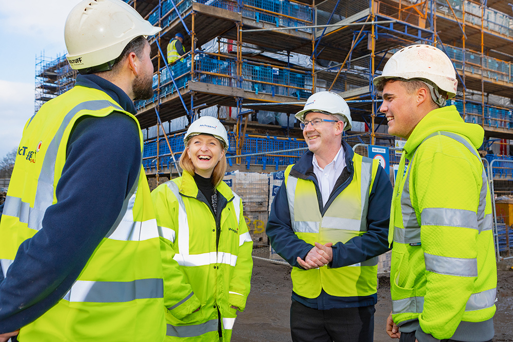 Three men and one women all wearing high visibility jackets and hard hats with a building site in the background.