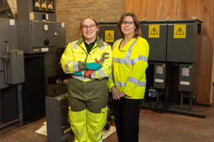 Two women, both wearing boiler suits, stand inside a control room.