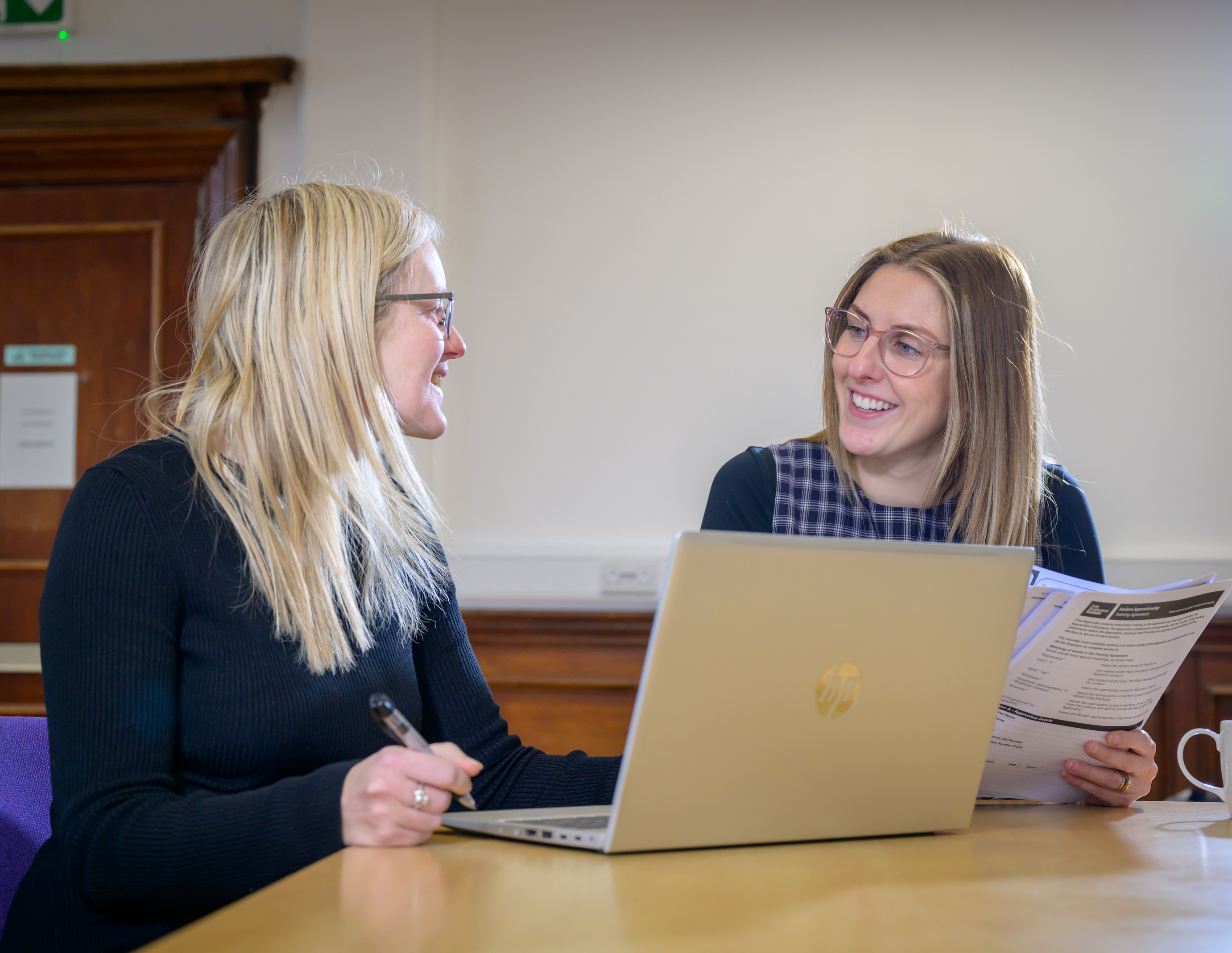 Two women sit at a desk with a laptop on it. They're smiling and talking to each other. One woman is holding a pen and the other woman is holding a piece of paper.